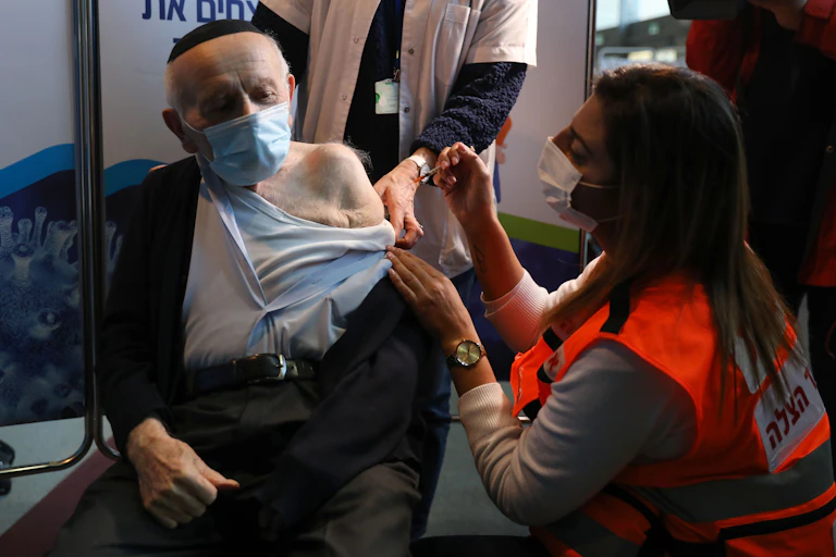 An elderly man wearing a yarmulke receives a vaccine from a young woman in a high-biz vest and mask.