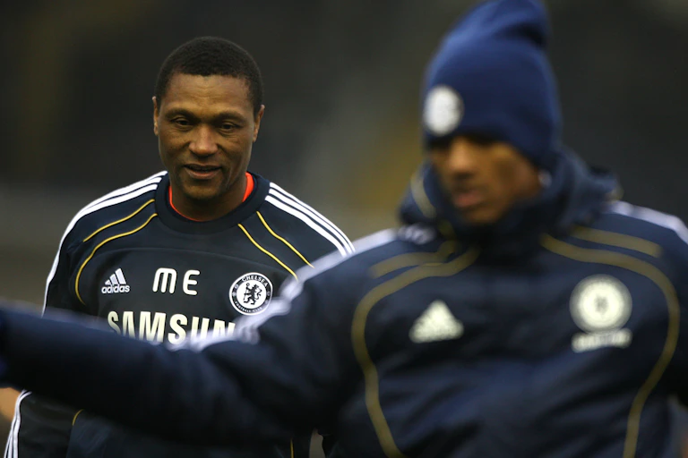 Two men, in focus an older male coach alongside an out of focus young man in a skull cap and tracksuit.