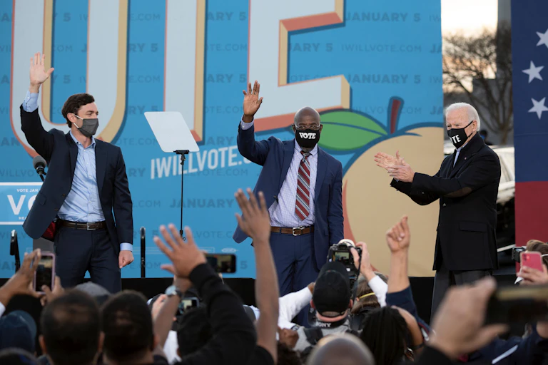 Ossoff and Warnock wave at a crowd of supporters, while Biden claps
