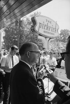 Black and white image of white man in glasses talking to press outside the Pickrick