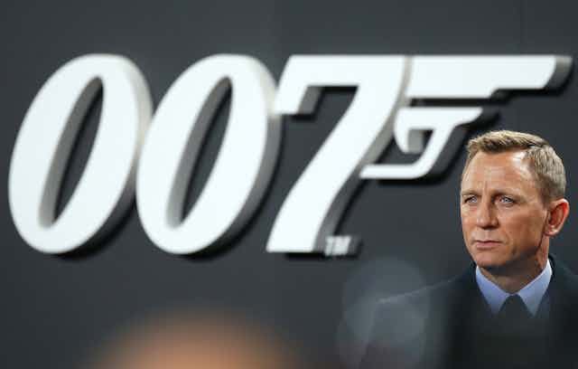 Daniel Craig wearing a suit and standing to the left of large 007 backdrop in black and white