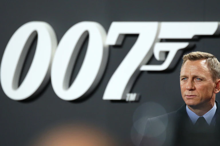 Daniel Craig wearing a suit and standing to the left of large 007 backdrop in black and white