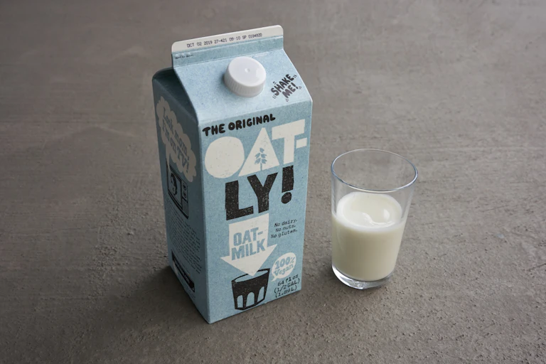 A carton of Oatly beside a glass of oat milk