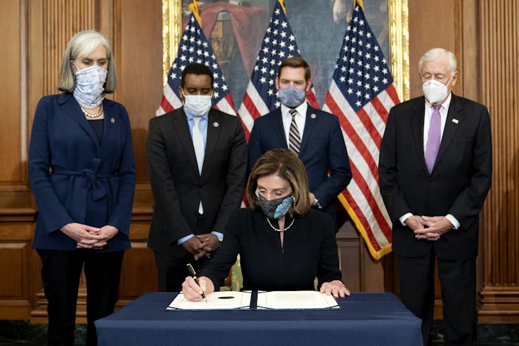 Pelosi signs a document with four people standing behind her, and American flags