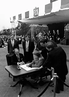 Queen Elizabeth sits at a small table and signs Canada's constitutional proclamation with Prime Minister Pierre Trudeau sitting at her side.