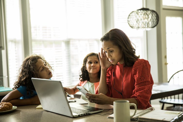 A woman tries to work on her computer while two young girls try to get her attention.