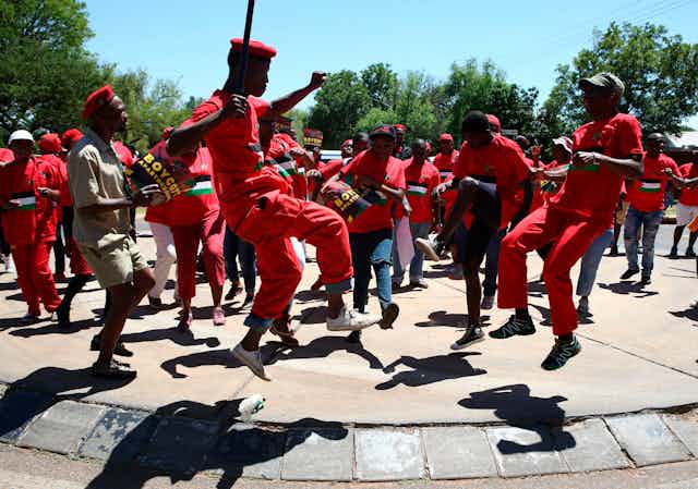 A group of men and women, dressed in red, form a circle, some leaping into the air, one crooked leg higher than the other, as they dance.