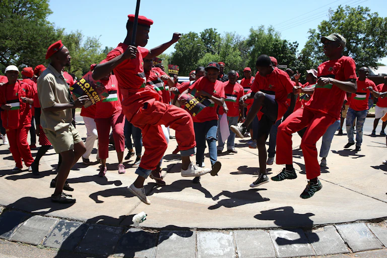 A group of men and women, dressed in red, form a circle, some leaping into the air, one crooked leg higher than the other, as they dance.