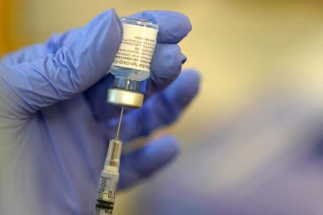 A gloved hand inserts a vial of the Pfizer vaccine into a syringe.