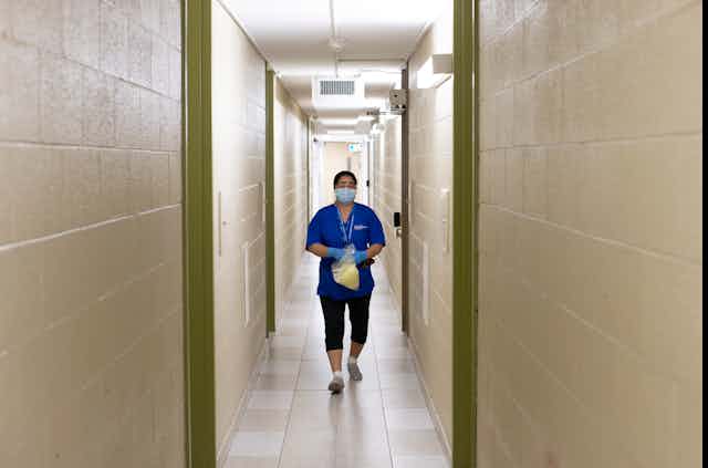 A woman in scrubs and a face mask in a long, narrow hallway