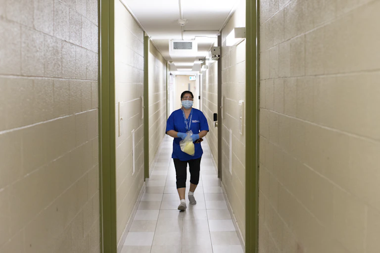 A woman in scrubs and a face mask in a long, narrow hallway