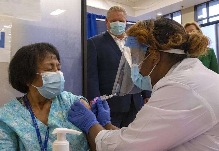 A woman in a mask gets an injection in her arm from another woman, while Premier Doug Ford stands in the background