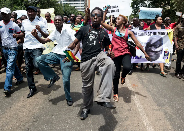 A group of protesters with banners; in the foreground a group appears to be marching in the same style, knees raised very high.