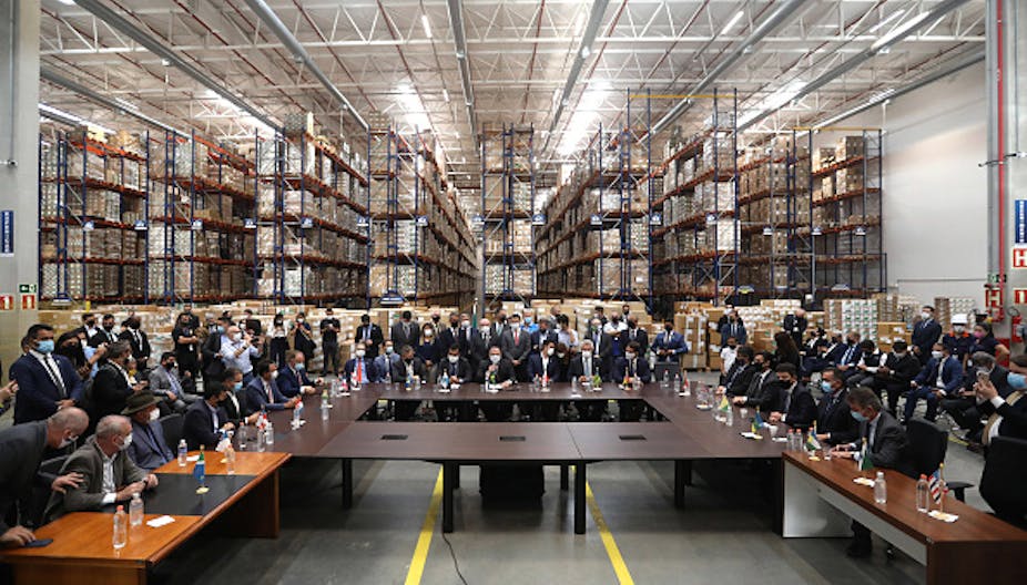 People wearing suits and face masks, seated and standing around conference table in a warehouse with shelves in the background