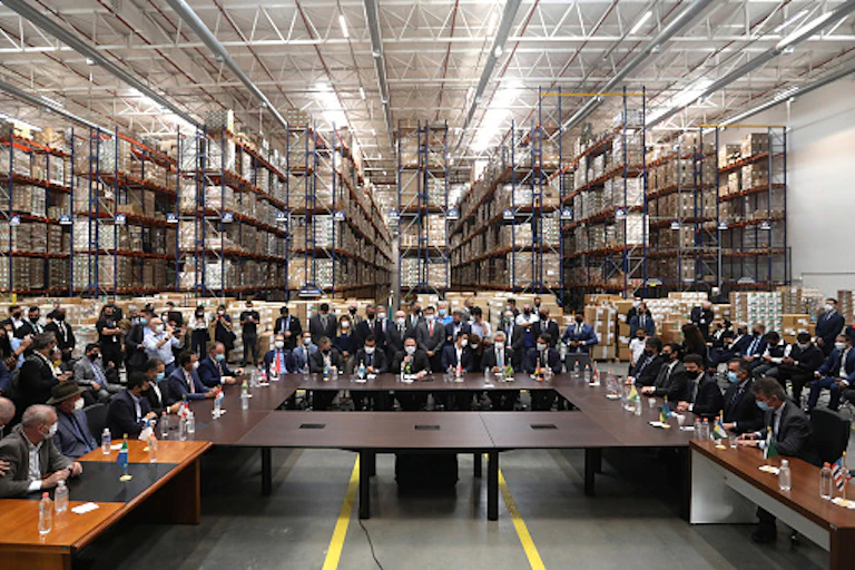 People wearing suits and face masks, seated and standing around conference table in a warehouse with shelves in the background
