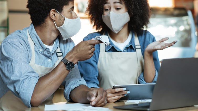 Financial problems in family small business during covid-19 pandemic. Sad young african american couple in aprons and protective masks gesturing and talking about bills and work with laptop in cafe