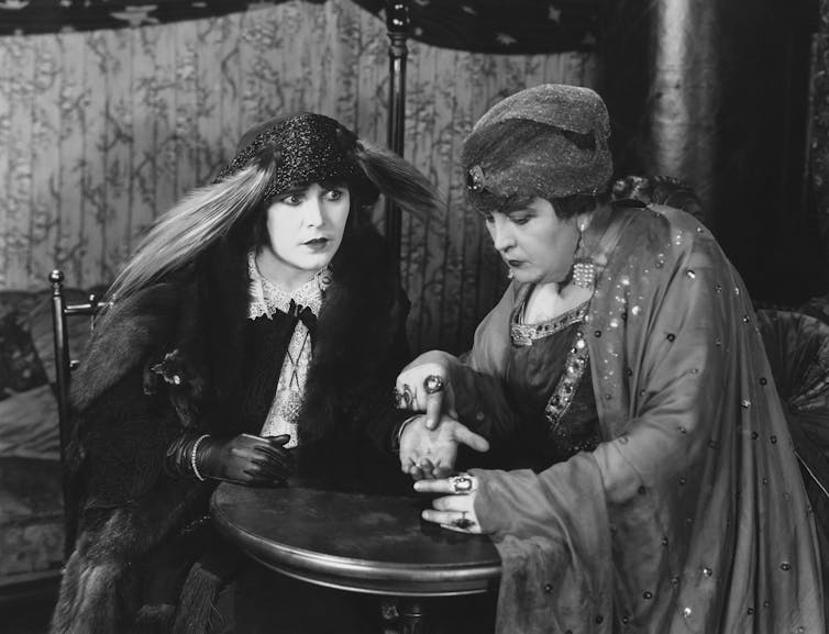 Two eccentrically-dressed women in a black and white photo, one reading the palm of the other