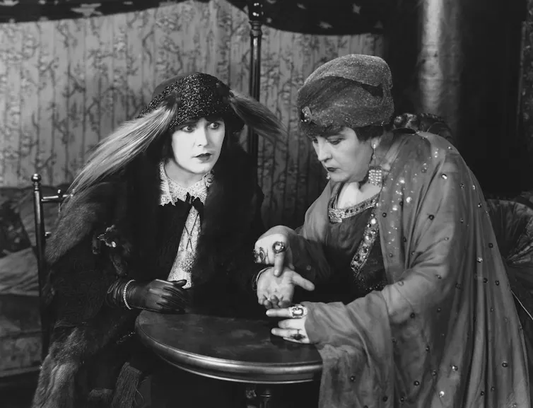 Two eccentrically-dressed women in a black and white photo, one reading the palm of the other