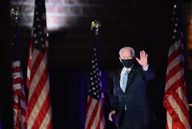 Biden walks in front of American flags while waving, in a black face mask