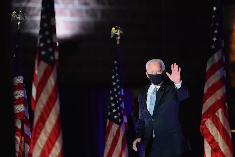 Biden walks in front of American flags while waving, in a black face mask