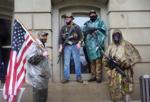 Four men carrying arms and an American flag in front of the Michigan State Capital building.