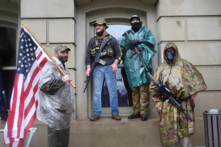 Four men carrying arms and an American flag in front of the Michigan State Capital building.