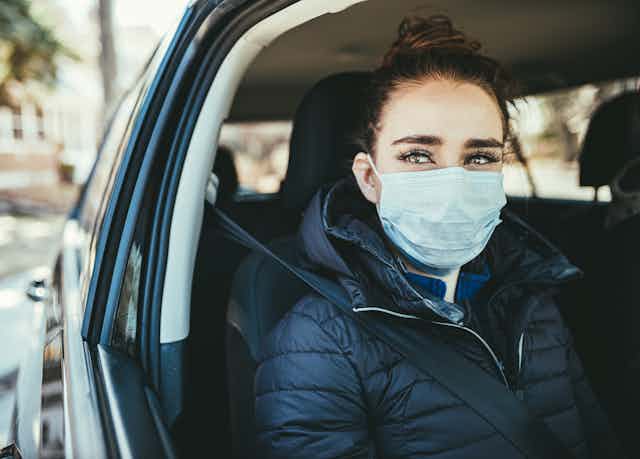 A woman wearing a medical mask looks out of an open driver's window of a car.