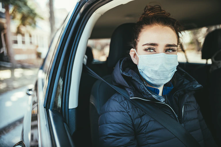 A woman wearing a medical mask looks out of an open driver's window of a car.