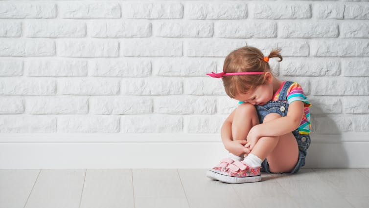 A little girl having a tantrum sits against a white brick wall.