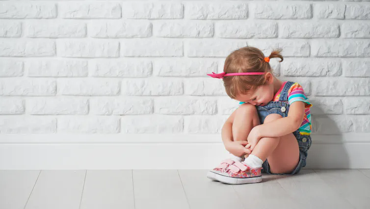 A little girl having a tantrum sits against a white brick wall.