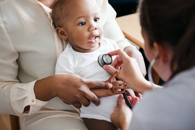 A doctor examines a toddler sitting on the mother's lap.