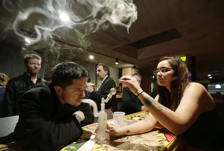 A man inhales marijuana smoke from a glass bong in a room with other people around.