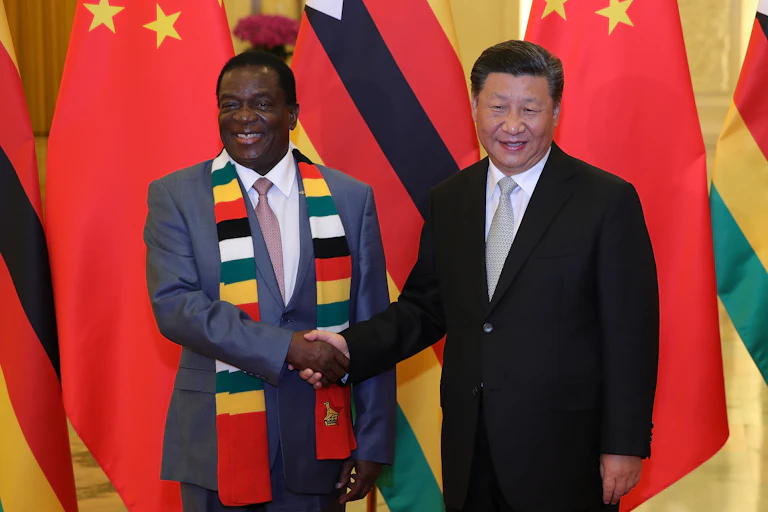 A man in a grey suit shakes with another in a black suit behind colourful flags