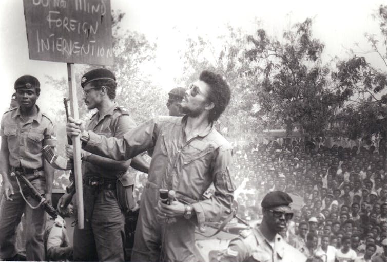 A man in an overall and shades reads a placard that says, 'Do Not Mind Foreign Intervention', a crowd of peoplein the background.