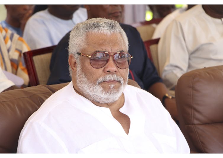 A greying man with a beard and sunglasses sits in a brown chair looking ahead intently.