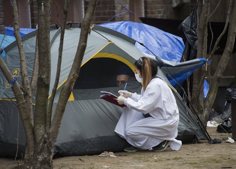 A woman wearing a mask and carrying a clipboard checks on a homeless person in a tent.