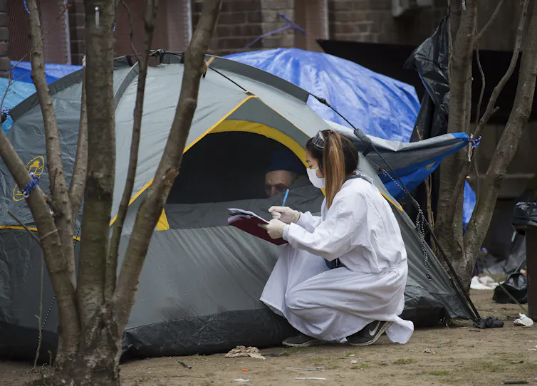A woman wearing a mask and carrying a clipboard checks on a homeless person in a tent.