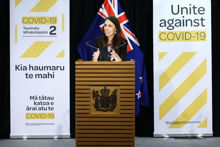 Jacinda Ardern standing behind podium between two COVID-19 health campaign banners