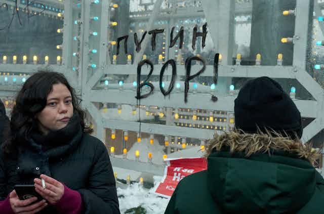 A woman holds a cigarette and a mobile phone in front of a message on a window in Cyrillic lettering which reads: 'Putin is a thief'.