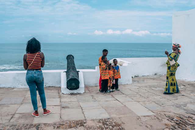 In front of a view of the sea from a fort with a cannon, a woman in traditional attire takes a photo of four children.