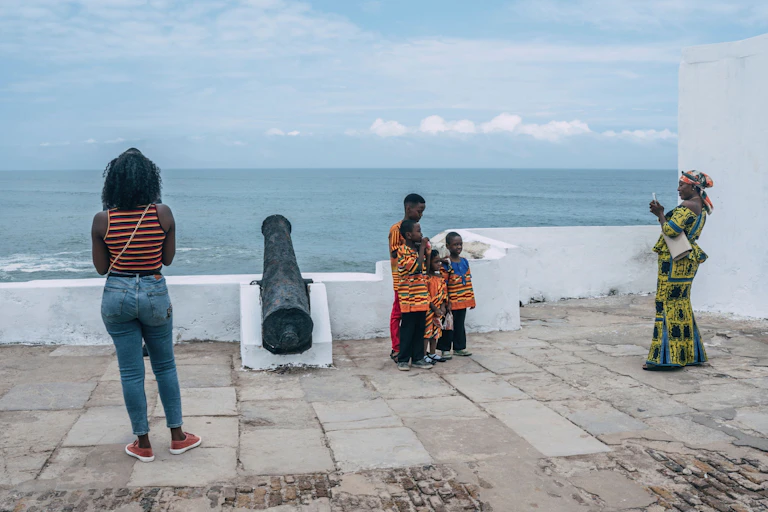 In front of a view of the sea from a fort with a cannon, a woman in traditional attire takes a photo of four children.