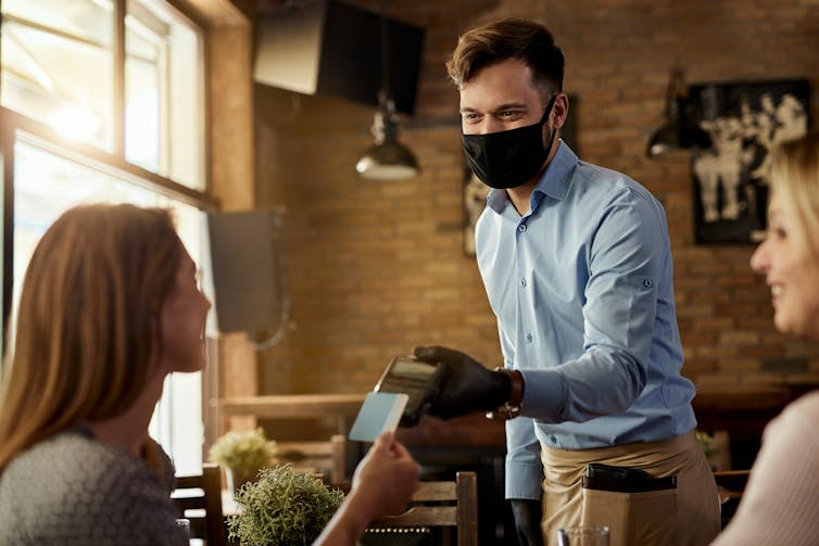 A man in a mask takes a contactless payment from a seated woman