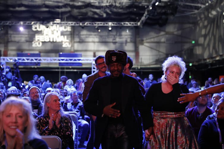 A glitzy room full of people, a man and a woman heading to stage, he in a hat and jacket, she in a full skirt and clutching her mouth in surprise.