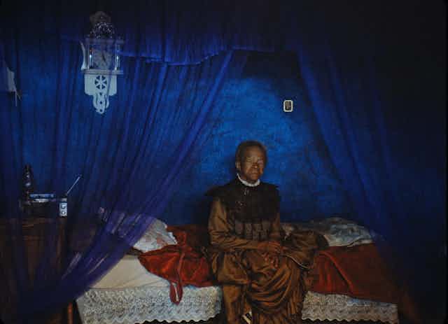 An elderly woman sits on a bed, dressed in a formal vintage gold dress against a backdrop of blue curtains and walls.
