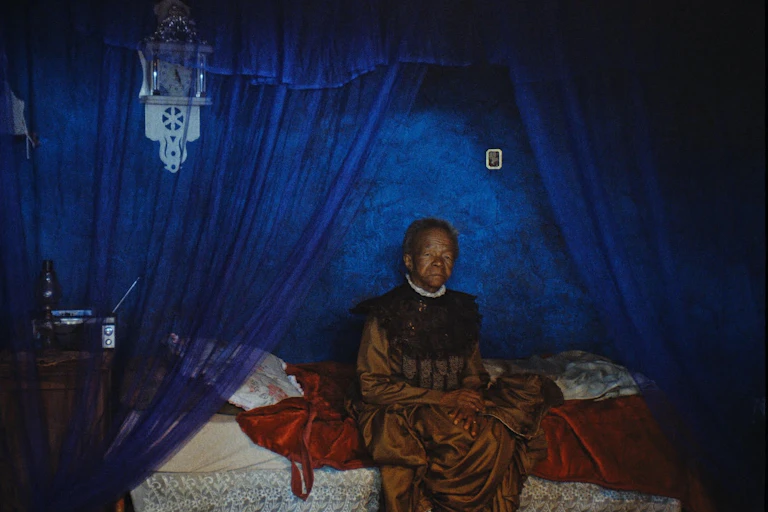 An elderly woman sits on a bed, dressed in a formal vintage gold dress against a backdrop of blue curtains and walls.