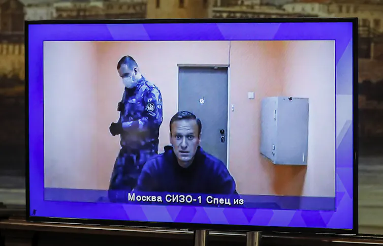 Video monitor showing Russian opposition activist Alexei Navalny in a jail cell with a Russian prison guard in the background.