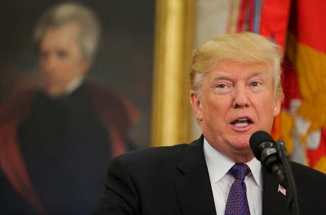 Donald Trump stands at a microphone in the Oval Office with a portrait of Andrew Jackson in the background.