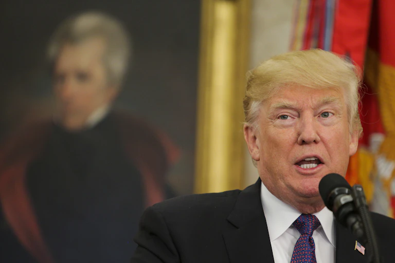 Donald Trump stands at a microphone in the Oval Office with a portrait of Andrew Jackson in the background.