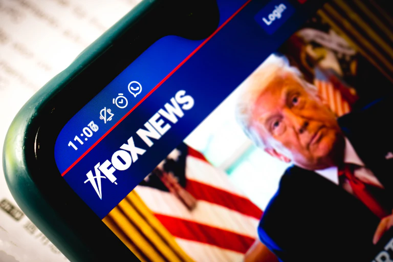 A close-up of a smartphone shows a Fox News article accompanied by an image of Donald Trump.