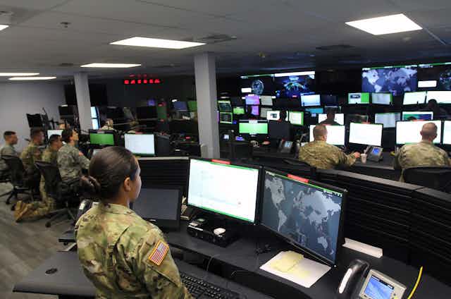 A woman in a military uniform looks at two computer screens in a room with other soldiers looking at other computer screens